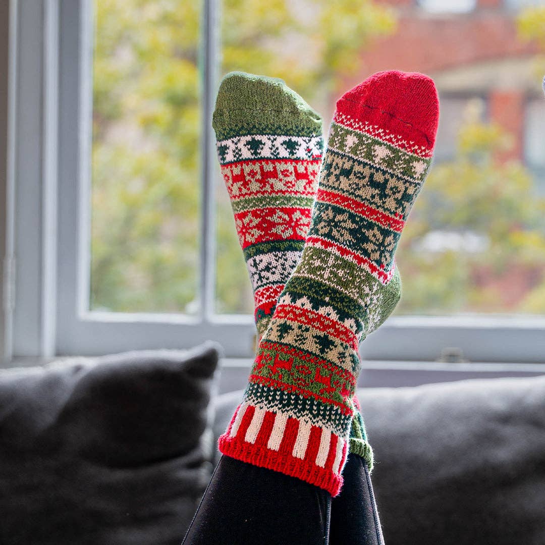 A pair of festive Christmas knit socks worn on feet resting on a dark gray couch in front of a window.