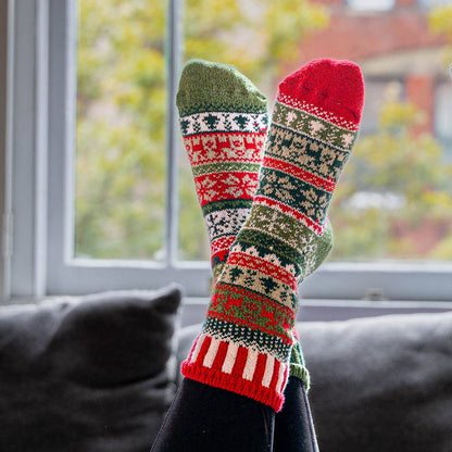 A pair of festive Christmas knit socks worn on feet resting on a dark gray couch in front of a window.