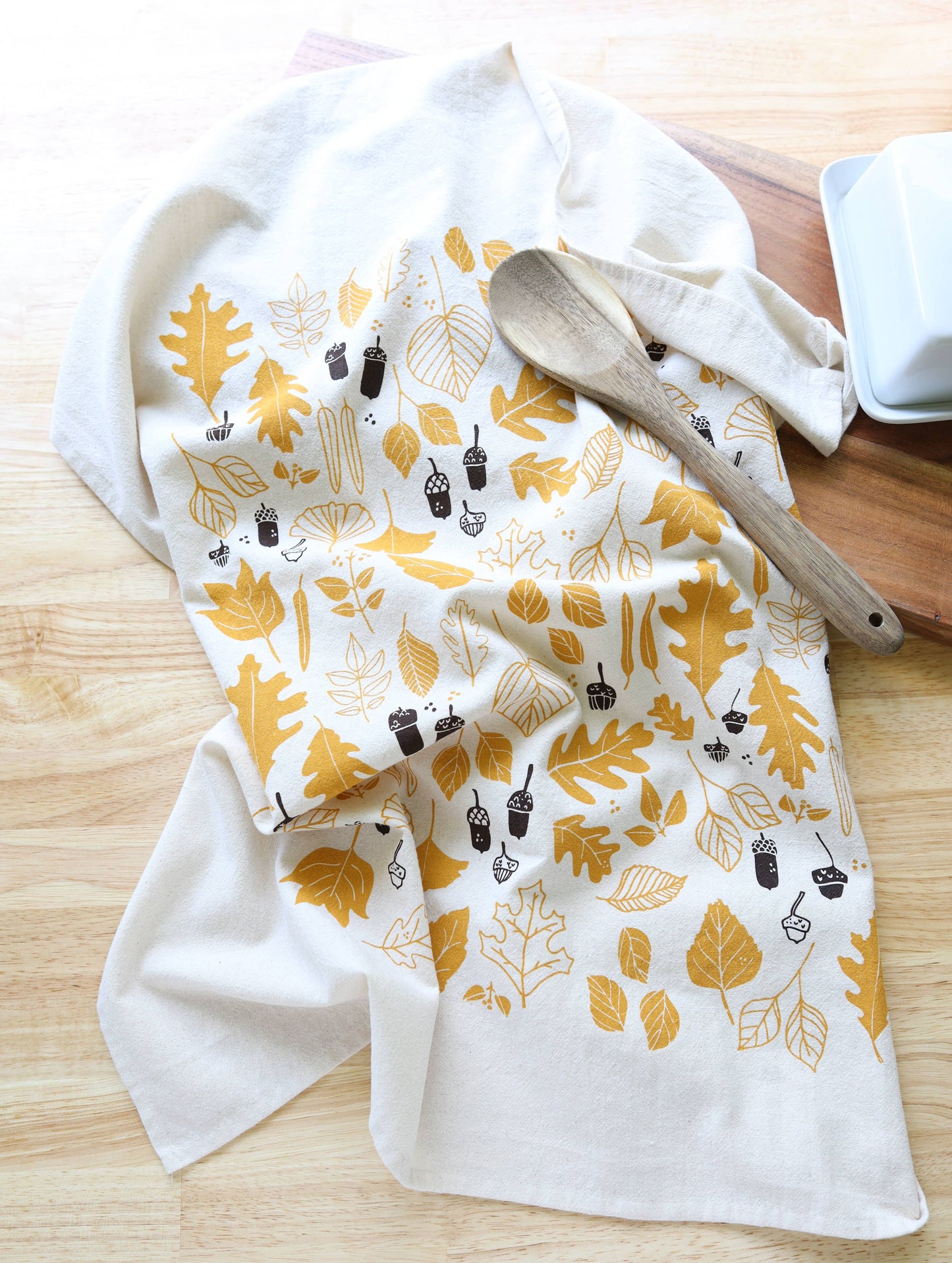 A white kitchen tea towel with a mustard yellow and dark brown autumn leaf pattern, styled on a light wood surface with a wooden spoon and a white dish.