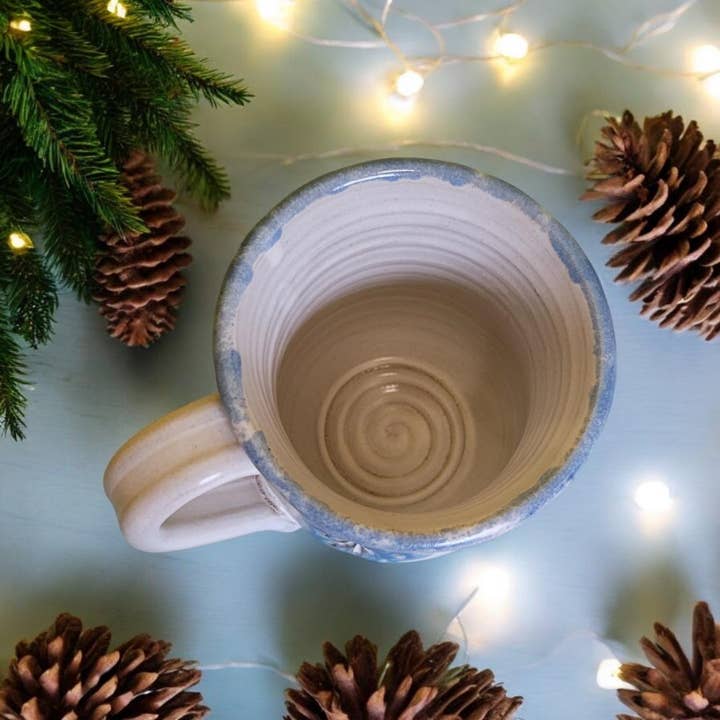 Ceramic mug with spiral design on a light blue surface surrounded by pine cones and string lights.