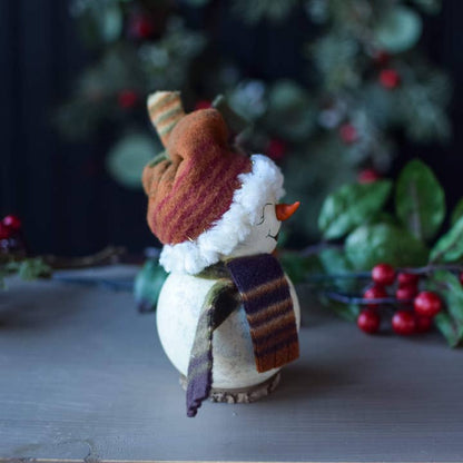 Decorative snowman figurine with a striped scarf and tree design, set against a blurred green and red background.