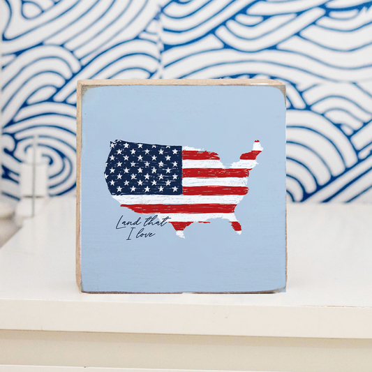 Small wooden sign with American flag design and 'Land that I love' text on a blue and white background.