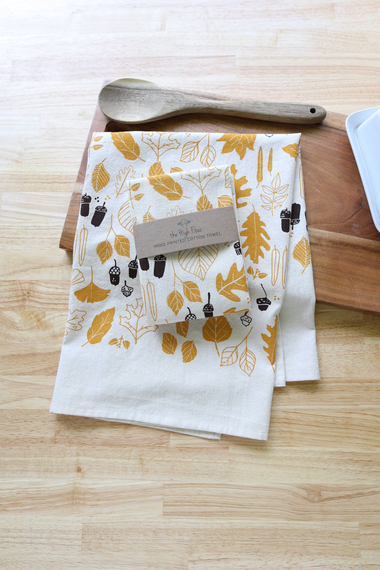 A folded white kitchen towel with a mustard yellow and dark brown leaf pattern, resting on a wooden cutting board with a wooden spoon. A paper tag is attached to the towel, which is placed on a light wood tabletop.
