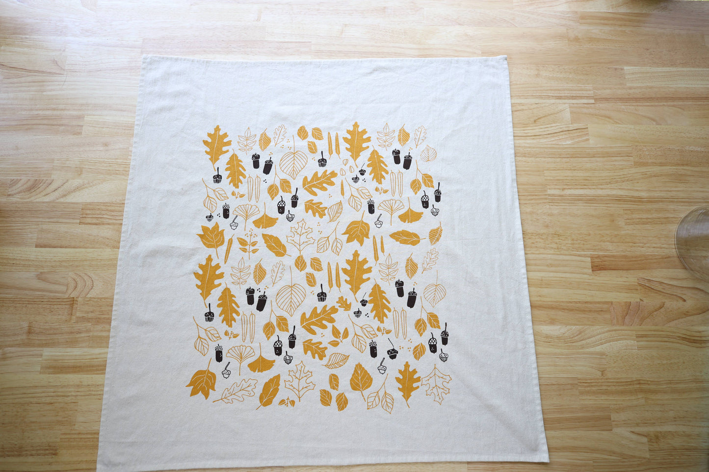A white tea towel or napkin laid flat on a light wood table. It features a printed pattern of autumn leaves and acorns in mustard yellow and dark brown.