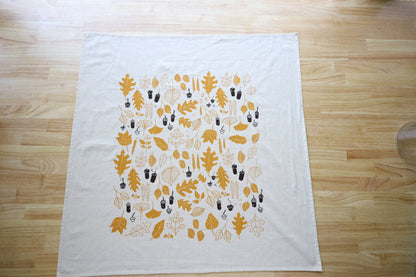 A white tea towel or napkin laid flat on a light wood table. It features a printed pattern of autumn leaves and acorns in mustard yellow and dark brown.