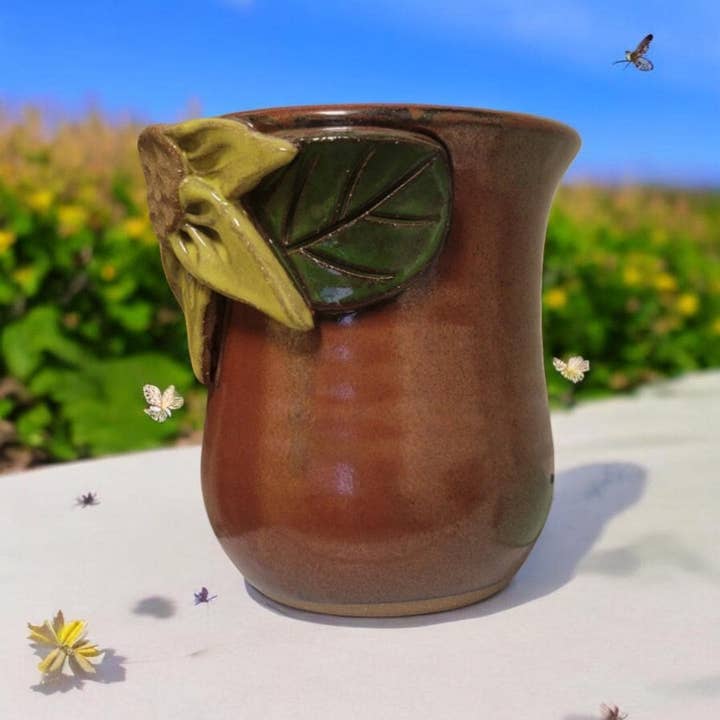 Brown ceramic mug with sunflower and leaf design on a white surface with a natural background