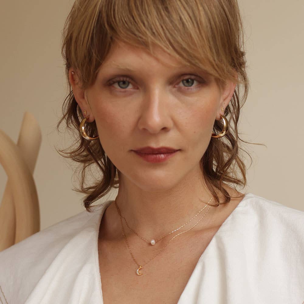 Woman wearing gold earrings and necklaces against a neutral background.