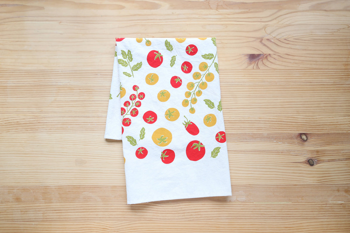 A white kitchen towel with a colorful print of red and yellow tomatoes and green leaves, displayed on a light wood grain surface.