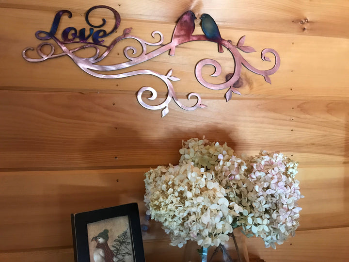 Decorative metal wall art with 'Love' and birds on a wooden wall, accompanied by a small framed picture and flowers.