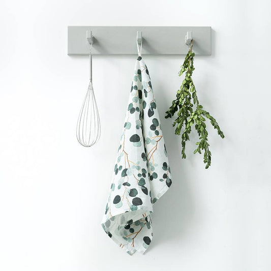 Gray wall hook with a whisk, green leaf print towel, and greenery on a white background