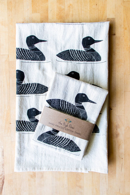 A natural beige cotton tea towel with a black Common Loon bird print, displayed on a wooden surface. The towel appears soft and absorbent.