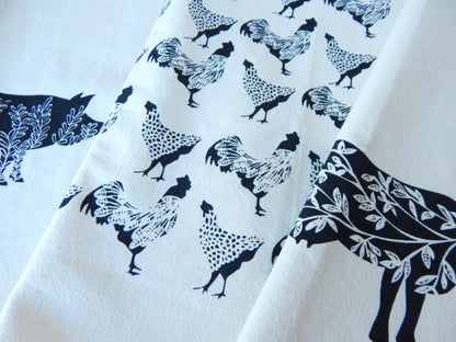 Close-up of a cotton tea towel with black printed chicken patterns and a larger black cow illustration on a white background.