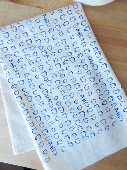 A folded beige cotton tea towel with blue printed teacup patterns on it, displayed on a wooden surface.