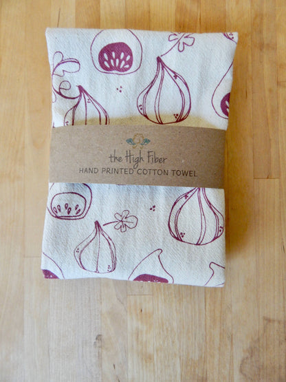 A beige cotton tea towel with purple fig prints, wrapped with a brown band displaying the brand name 'The High Fiber'.