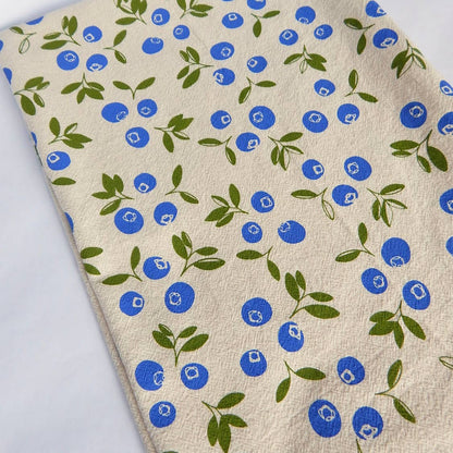 A beige cotton tea towel with a blueberry print design in a natural color palette.