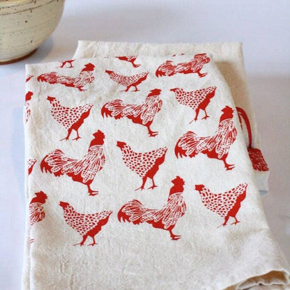 A folded beige cotton tea towel with red printed chickens