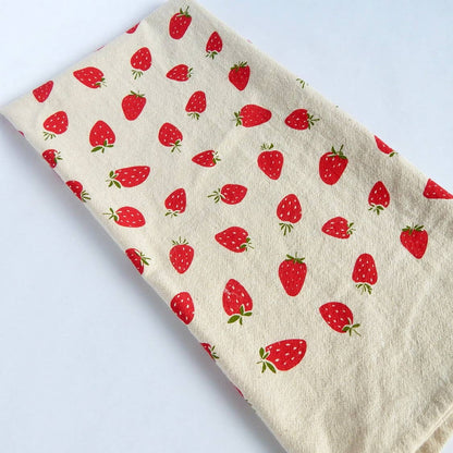 A cotton tea towel with a printed pattern of red strawberries on a natural beige background.