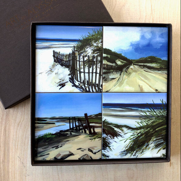 A set of four ceramic tile coasters with cork backing, each featuring a different image of a beach scene, displayed in a dark brown gift box.