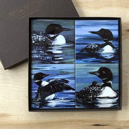 A set of four ceramic tile coasters with cork backing, each featuring an image of a loon bird in various poses, displayed in a black gift box with 'Art by Alyssa' logo.