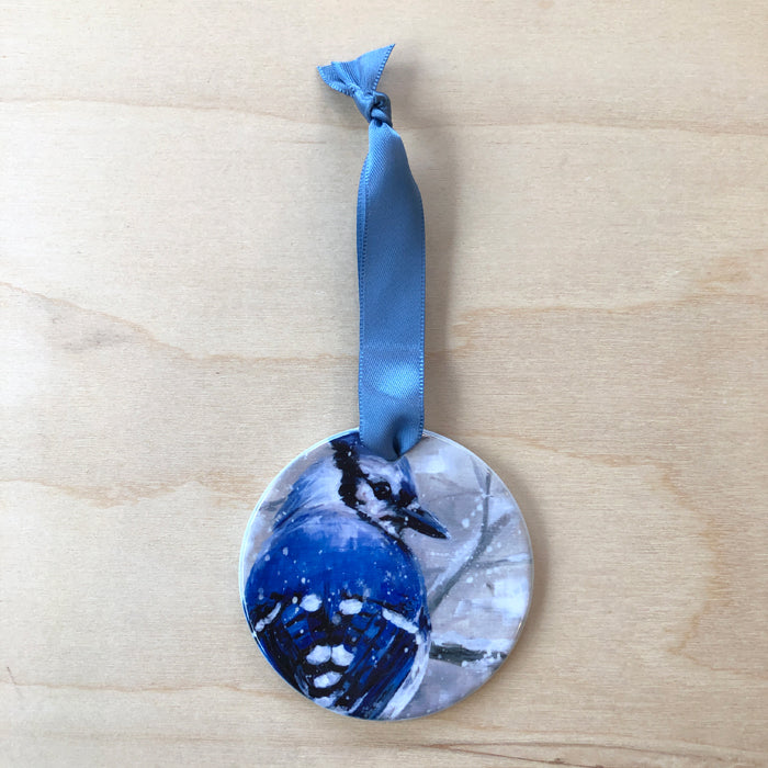 A double-sided porcelain ornament featuring an image of a bluejay with a blue satin ribbon attached to the top.