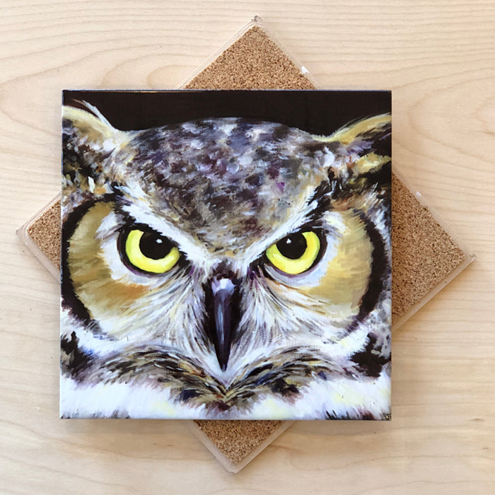 A square trivet with a cork backing featuring an image of a great horned owl with yellow eyes.