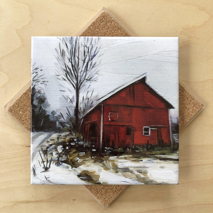 A square ceramic trivet with an image of a red barn in winter, surrounded by a white snowy landscape and a leafless tree to the left, placed on a cork backing.