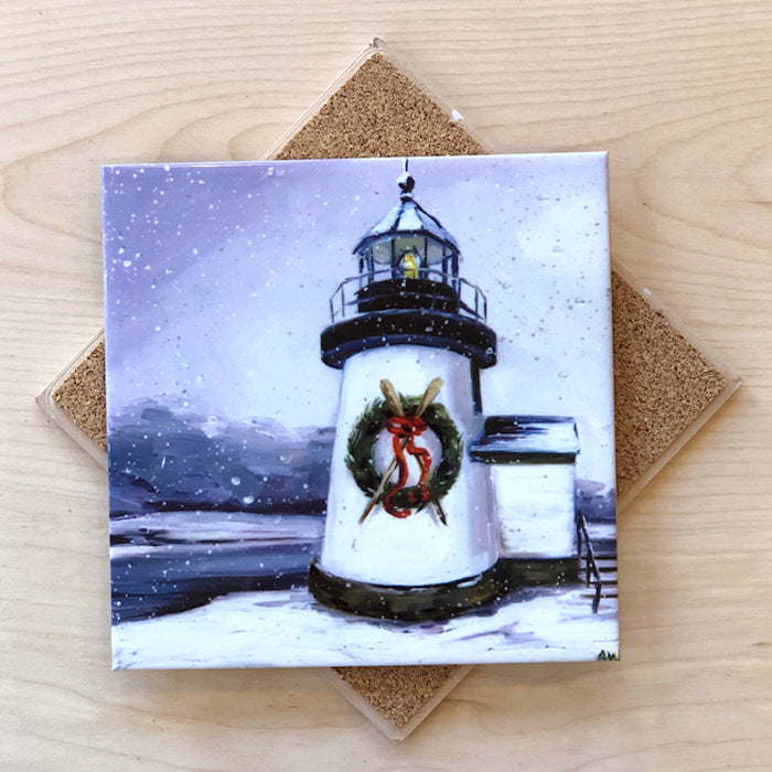 A square ceramic trivet with an image of a lighthouse adorned with a Christmas wreath, placed on a wooden surface with cork backing.