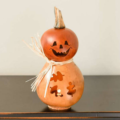 A decorative gourd lantern shaped like a jack-o'-lantern with a carved face, small leaf cutouts, a straw bow, and a short stem, shown on a dark tabletop.