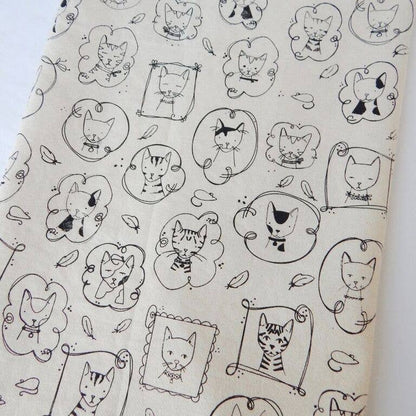 A beige cotton tea towel with a pattern of various cartoon cat faces in black ink.