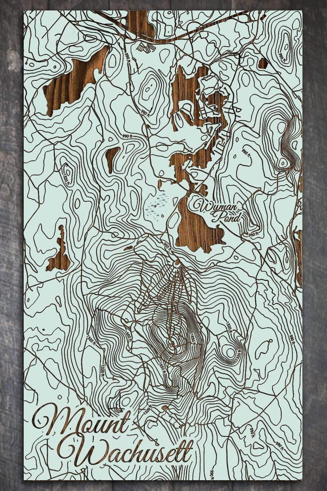 A wooden map of Mount Wachusett featuring topographical lines, roads, and a nearby pond, engraved in sea green color on a pine panel.